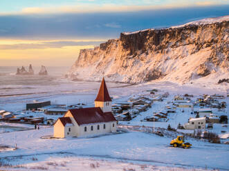 Der Sonnenuntergang wirft ein warmes Licht über ein kleines isländisches Dorf mit einer Kirche zwischen schneebedeckten Klippen und dem Meer - ADSF52435