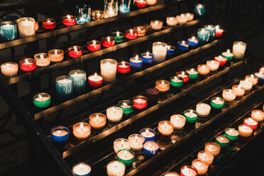 Reihen von bunten Votivkerzen, die in einer schwach beleuchteten Kirche in Bayonne sanft auf gestuften Ständern leuchten und eine Atmosphäre der Ehrfurcht und Meditation schaffen - ADSF52498