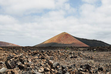 Ein markanter Vulkankegel erhebt sich über einem zerklüfteten Feld aus Lavagestein unter einem teilweise bewölkten Himmel auf Lanzarote - ADSF52500