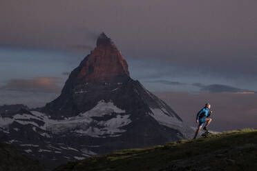 A trail runner treks across an alpine landscape against the striking silhouette of a majestic mountain at dusk, showcasing nature's grandeur and the spirit of adventure - ADSF52515
