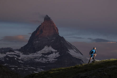 Ein Trailrunner wandert durch eine alpine Landschaft vor der beeindruckenden Silhouette eines majestätischen Berges in der Abenddämmerung, die die Erhabenheit der Natur und den Geist des Abenteuers zeigt - ADSF52515