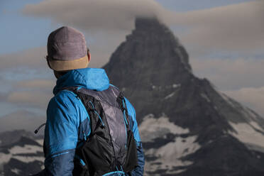 Ein Wanderer in einer blauen Jacke betrachtet einen majestätischen, wolkenverhangenen Berggipfel - ADSF52517