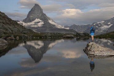 Eine einsame Gestalt steht auf einem Felsen an einem ruhigen Bergsee und spiegelt sich im sanften Schein des Sonnenuntergangs auf dem ruhigen Wasser - ADSF52518