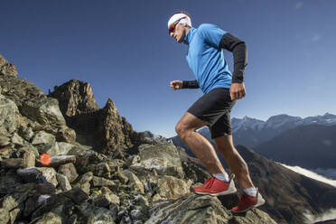 Focused male runner in sportswear powerfully ascending a rocky alpine path with jagged peaks in the background - ADSF52520