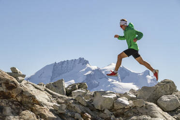 Full body of runner in a green jacket leaping between rocks with snowy mountains in the background under a clear blue sky - ADSF52526