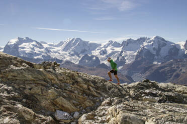 Ein einsamer Läufer in farbenfroher Sportkleidung stapft durch eine zerklüftete Berglandschaft mit schneebedeckten Gipfeln in der Ferne - ADSF52527