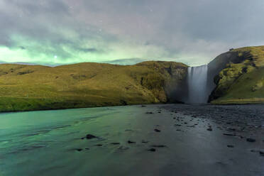 Ein himmlischer Blick auf den Wasserfall Skogafoss, der unter einem sternenklaren Dämmerungshimmel eine üppig grüne Klippe hinabstürzt, mit ruhigem Wasser im Vordergrund. Island - ADSF52539