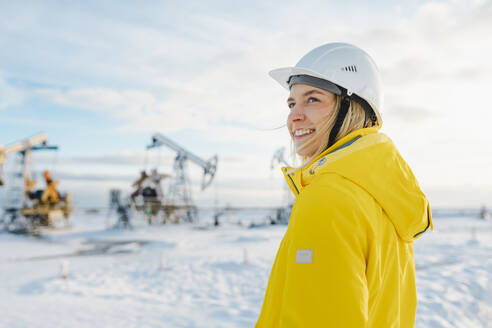 Lächelnder junger Ingenieur mit Schutzhelm auf einem Ölförderfeld im Winter - OLRF00104