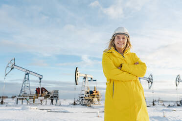 Lächelnder junger Ingenieur mit verschränkten Armen auf einem Ölförderfeld im Winter - OLRF00105