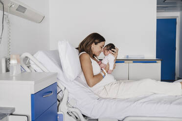 Mother sitting on bed and embracing baby daughter in hospital - EBBF08276