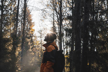 Side view of unrecognizable bearded male traveler in warm clothes and hat with backpack standing and looking up in autumn forest at Stockholm National Park on sunny day - ADSF52558