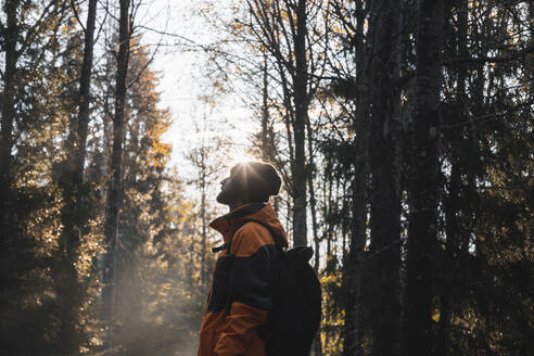 Seitenansicht eines nicht erkennbaren bärtigen männlichen Reisenden in warmer Kleidung und mit Hut, der mit einem Rucksack im Herbstwald des Stockholmer Nationalparks steht und nach oben blickt, an einem sonnigen Tag - ADSF52558