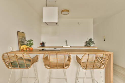 Induktion und Obst auf einer Holzinsel mit leeren Stühlen in einer modernen Küche einer luxuriösen Wohnung mit weißen Wänden - ADSF52564