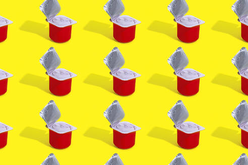 A repetitive pattern of red cups of strawberry yogurt, each with its foil lid opened, arranged neatly on a bright yellow background - ADSF52575