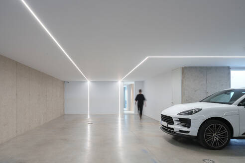 Unkenntlich gemachte Person in einer modernen Garage mit integrierter LED-Beleuchtung, poliertem Betonboden und einem geparkten Luxusauto - ADSF52583