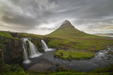 Üppige grüne Landschaft mit einem markanten Berggipfel, der sich über zwei Wasserfälle unter einem dramatischen Himmel in Island erhebt - ADSF52584