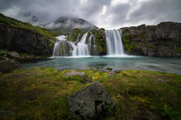 Üppige grüne Landschaft mit einem markanten Berggipfel, der sich über zwei Wasserfälle unter einem dramatischen Himmel in Island erhebt - ADSF52585