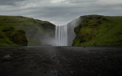 Mächtiger Wasserfall, der eine üppige grüne Klippe hinunter in ein nebliges Becken mit dunklen Kieselsteinen im Vordergrund in Island stürzt - ADSF52589
