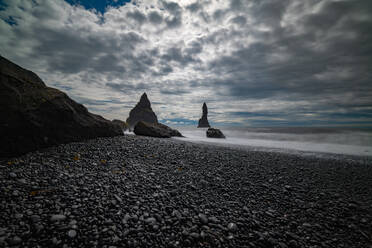 Zerklüftete Felsen ragen aus dem nebligen Meer vor dem Kiesstrand eines dramatischen isländischen Strandes unter einem wolkenverhangenen Himmel - ADSF52591