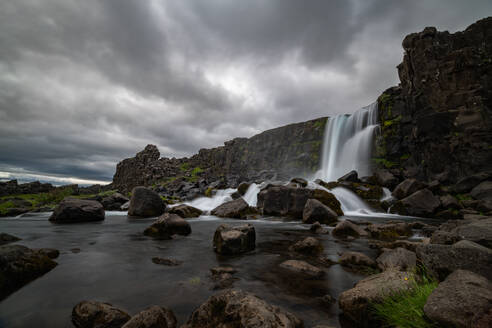 Ein dramatischer Wasserfall, der unter einem stürmischen Himmel in der zerklüfteten Landschaft Islands über alte Basaltsäulen stürzt - ADSF52593