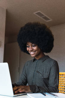Glückliche afroamerikanische Freiberuflerin mit stilvoller Afrofrisur, die ein modernes Netbook durchsucht, während sie aus der Ferne an einem Tisch in einem hellen Raum arbeitet - ADSF52596