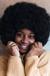 Ausschnitt einer fröhlichen jungen Afroamerikanerin mit Afrofrisur und geschlossenen Augen, die ihren Pullover anfasst, während sie an einer grauen Wand steht - ADSF52599