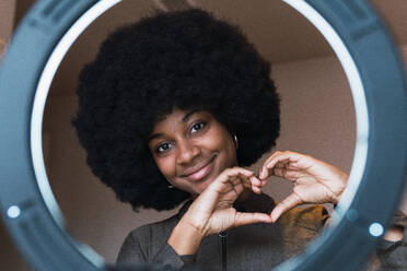 Cheerful African American female blogger with Afro hairstyle making heart gesture with hands and looking at camera through ring lamp - ADSF52602