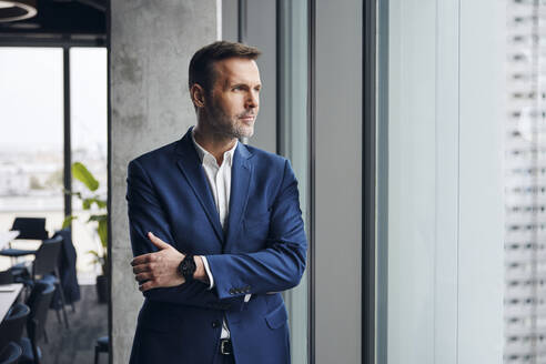 Thoughtful mature businessman standing with arms crossed near window in office - BSZF02376