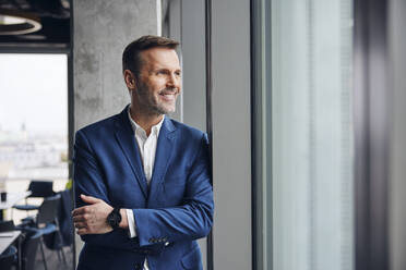 Smiling businessman standing with arms crossed near window in office - BSZF02377