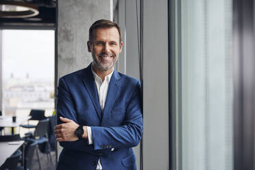 Happy businessman standing with arms crossed near window in office - BSZF02378