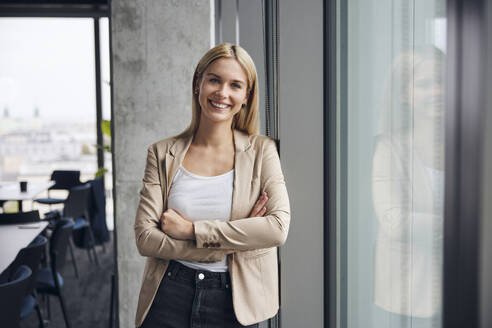 Lächelnde Geschäftsfrau, die mit verschränkten Armen in der Nähe des Fensters im Büro steht - BSZF02379