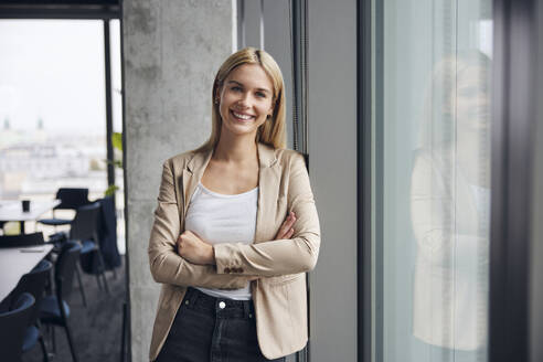 Smiling businesswoman standing with arms crossed near window in office - BSZF02379