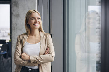 Happy businesswoman standing with arms crossed near window in office - BSZF02381