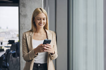 Glückliche Geschäftsfrau mit Smartphone am Fenster im Büro - BSZF02382