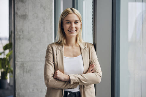 Happy businesswoman standing near window in office - BSZF02383