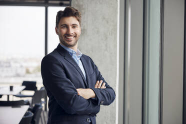 Happy young businessman standing with arms crossed in office - BSZF02385