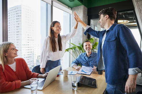 Happy businesswoman giving high-five to businessman near colleagues sitting at desk - BSZF02390