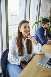 Smiling businesswoman sitting with laptop near colleague at desk - BSZF02398