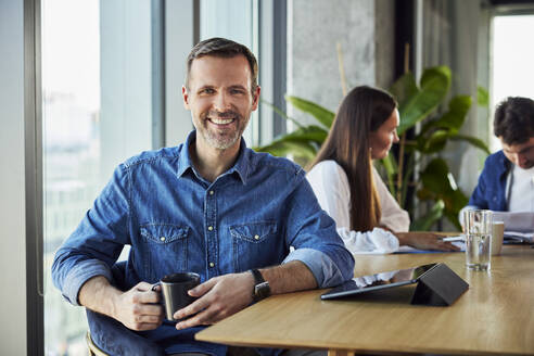 Happy businessman sitting with tablet PC and coffee cup near colleagues - BSZF02406