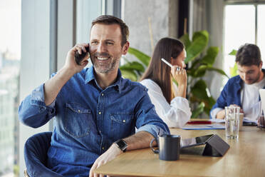 Glücklicher Geschäftsmann, der am Schreibtisch mit seinem Smartphone telefoniert - BSZF02407