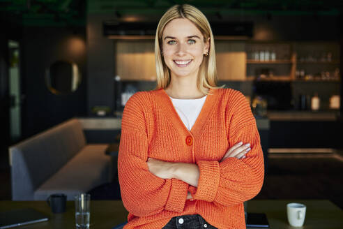 Happy blond businesswoman standing with arms crossed in modern office - BSZF02450