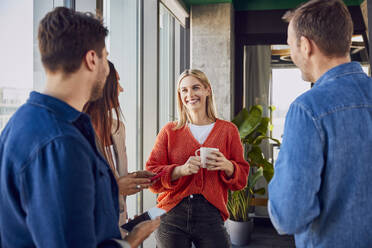 Happy businesswoman holding coffee cup and discussing with colleagues in office - BSZF02458