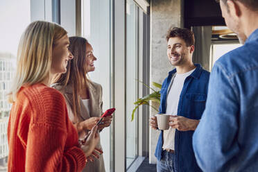 Smiling businessman holding coffee cup and talking to colleagues in office - BSZF02459