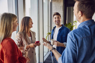 Happy businessman holding coffee cup and talking to colleagues in office - BSZF02460