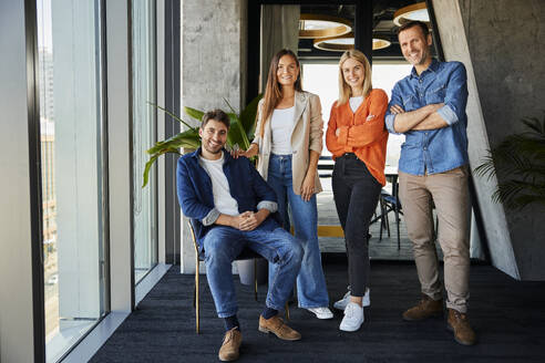 Smiling businessman sitting on chair with colleagues standing in office - BSZF02466