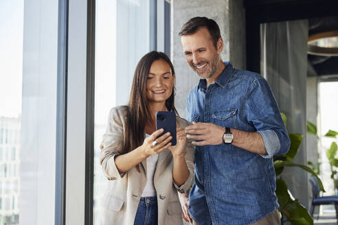 Lächelnde Geschäftsfrau, die mit einem Kollegen im Büro ein Smartphone benutzt - BSZF02472