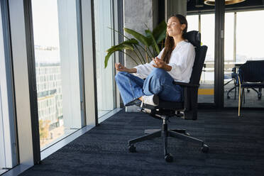 Smiling businesswoman practicing meditation on office chair - BSZF02488