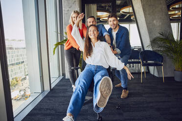 Happy colleagues pushing businesswoman sitting on chair and having fun in office - BSZF02489