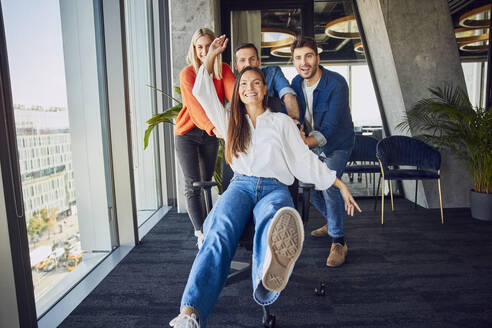 Happy colleagues pushing businesswoman sitting on chair and having fun in office - BSZF02489