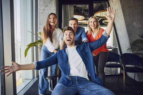 Happy colleagues pushing businessman sitting on chair and having fun in office - BSZF02491